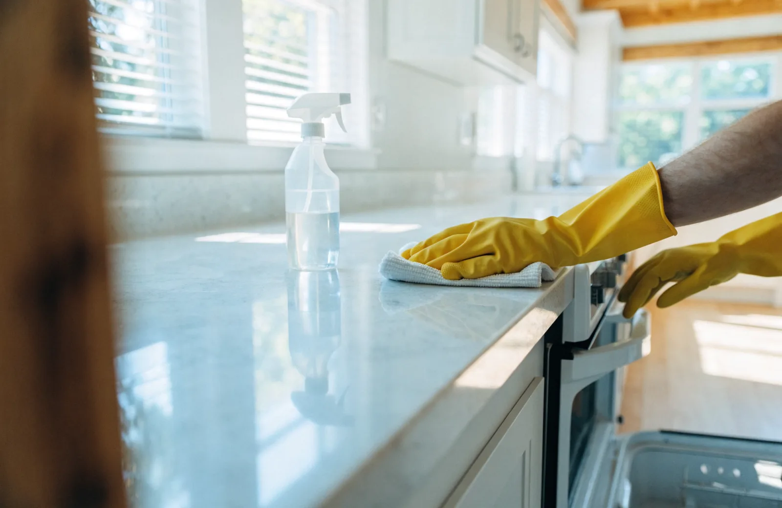 Gloved hands wiping a white kitchen countertop with a spotless open oven in the background